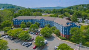 Aerial Exterior of Centennial Court, trees planted throughout the property, parking in front, sprawling hills in the distance, building painted blue, beautifully groomed grounds, lush foliage, photo taken on a sunny day.