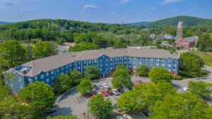 Aerial Exterior of Centennial Court, trees planted throughout the property, parking in front, sprawling hills in the distance, building painted blue, beautifully groomed grounds, lush foliage, photo taken on a sunny day.