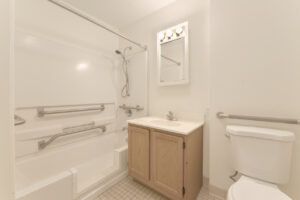 Interior Unit bathroom, Light brown cabinetry, vanity mirror, handicap accessible shower, with extra railings, tile floor.